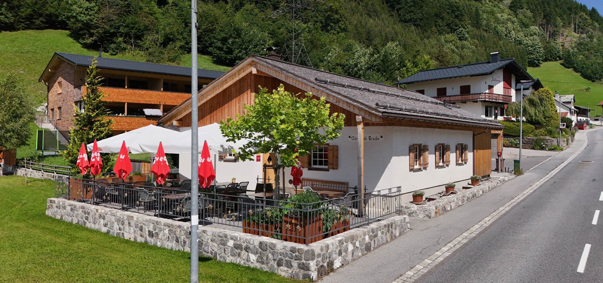 Ein rustikales Gasthaus mit einem Außenbereich, der von einem Geländer umgeben ist. Rote Sonnenschirme stehen auf der Terrasse, um Gästen Schatten zu spenden. Im Hintergrund sind grüne Hügel und weitere Häuser zu sehen.