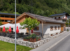 Eine rustikale Gaststätte mit hellem Verputz und Holzakzenten. Vor dem Gebäude stehen Tische unter Markisen, umgeben von roten Schirmen. Im Hintergrund sind Bäume und weitere Häuser sichtbar, die die alpine Umgebung ergänzen.