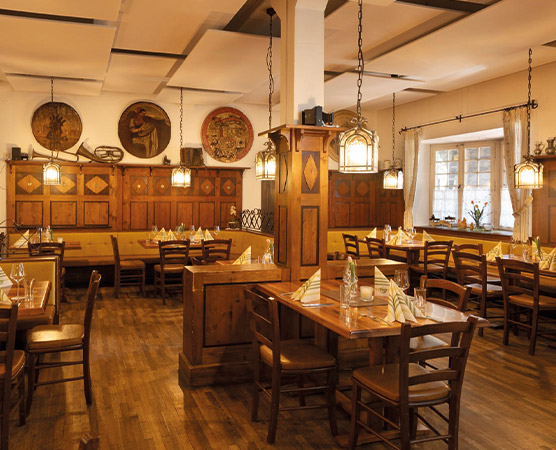 Ein gemütliches Restaurant mit Holzmöbeln. Mehrere Tische sind eingedeckt, mit Servietten gefaltet. An den Wänden hängen runde Wandmalereien. Sanftes Licht strömt von hängenden Lampen, und große Fenster lassen Tageslicht herein. Der Boden ist aus Holz, was eine warme Atmosphäre schafft.