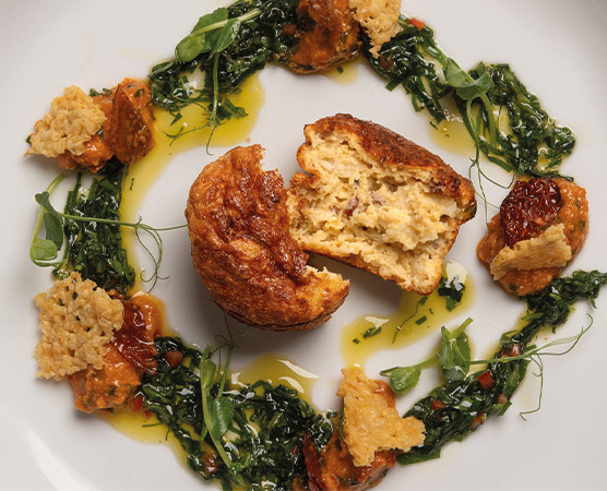 Ein kunstvoll angerichteter Teller mit einem aufgeschnittenen Muffin in der Mitte, umgeben von grünen Blättern und kleinen Portionen von Tomatenpüree und knusprigem Brot. Ein Spritzer Olivenöl ergänzt die Präsentation.