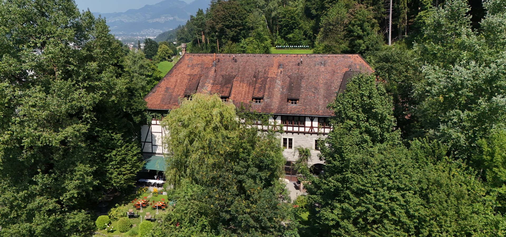 Ein großes, traditionelles Gebäude mit einem schrägen, roten Dach ist von dichtem, grünem Laub umgeben. Vor dem Gebäude sind Sitzgelegenheiten im Freien und ein gepflegter Gartenbereich sichtbar. Im Hintergrund erheben sich sanfte Hügel und eine Berglandschaft.