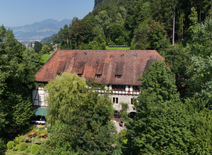 Das Bild zeigt ein traditionsreiches, zweigeschossiges Gebäude mit einem steilen, roten-satteldach, umgeben von üppigem Grün und Bäumen. Im Vordergrund sind Blumenbeete zu sehen. Die Landschaft im Hintergrund deutet auf eine bergige Umgebung hin.