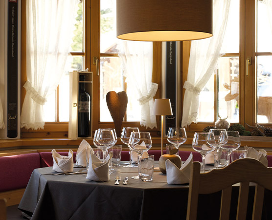 Ein elegant gedeckter Tisch in einem Restaurant, umgeben von hellen Fenstern und sanfter Beleuchtung. Der Tisch ist mit Gläsern, Servietten und Besteck geschmückt, unter einem großen Lampenschirm. Eine gemütliche Atmosphäre lädt zum Verweilen ein.