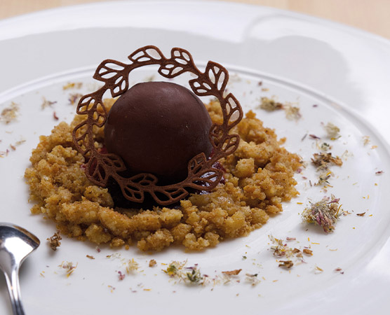 Ein eleganter Dessertteller mit einer Schokoladenkugel in der Mitte, umgeben von bröseligem Keksboden. Um die Kugel ist ein filigranes Schokoladenhäubchen angeordnet. Der Teller hat zusätzlich einige essbare Blütenstreu und einen Löffel auf der Seite.