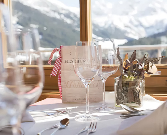Ein elegant eingedeckter Tisch mit mehreren Weingläsern, Besteck und einer kleinen Pflanze in einem Glas. Im Hintergrund sieht man die verschneiten Berge durch große Fenster. Eine Menükarte mit einer roten Schleife steht ebenfalls auf dem Tisch.