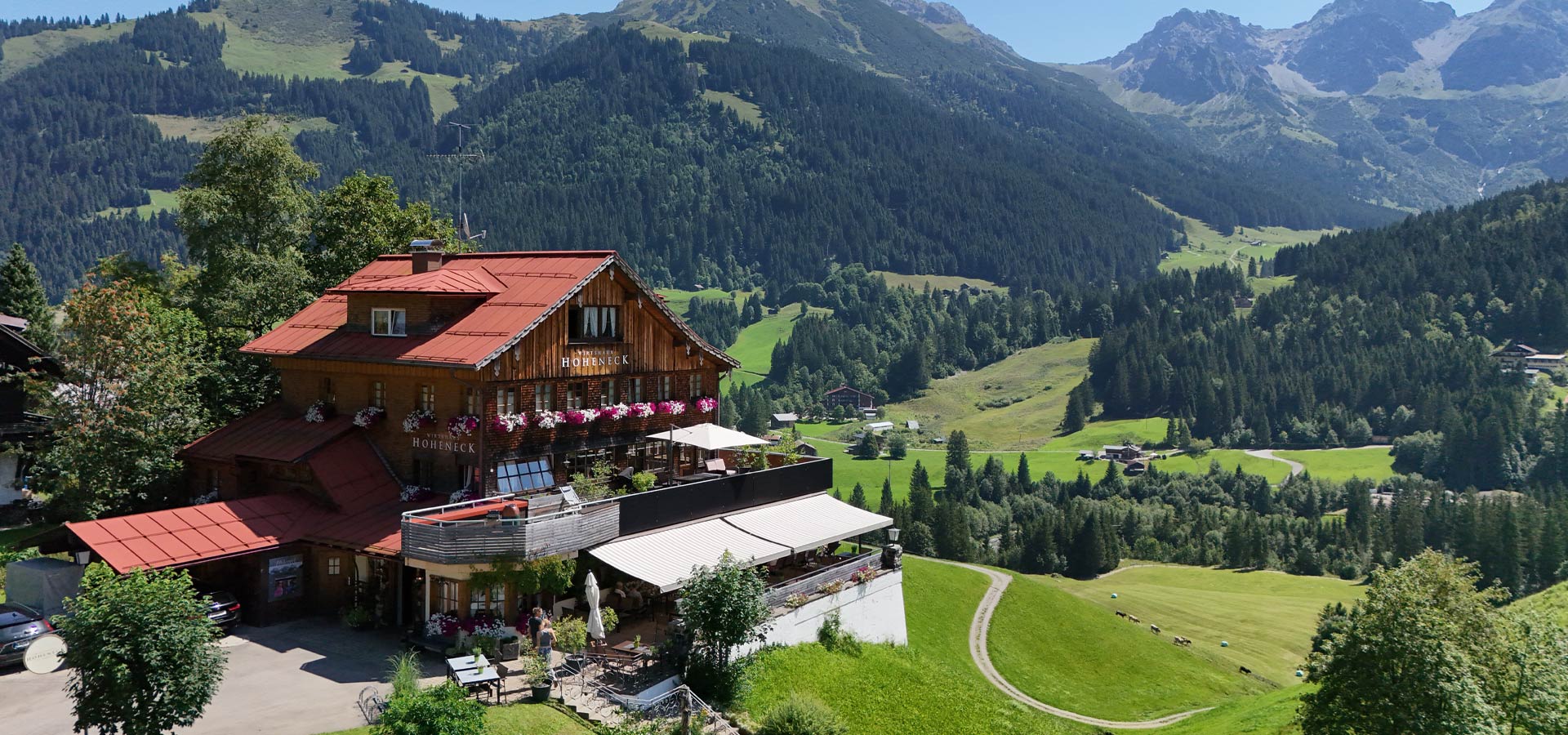 Ein traditionelles Holzhaus mit rotem Satteldach steht malerisch in einer grünen Landschaft. Umgeben von Bergen und Wäldern, erfährt es eine schöne Aussicht. Auf der Terrasse sind Tische und Stühle eingerichtet, und blühende Pflanzen verleihen dem Ort einen einladenden Charme.