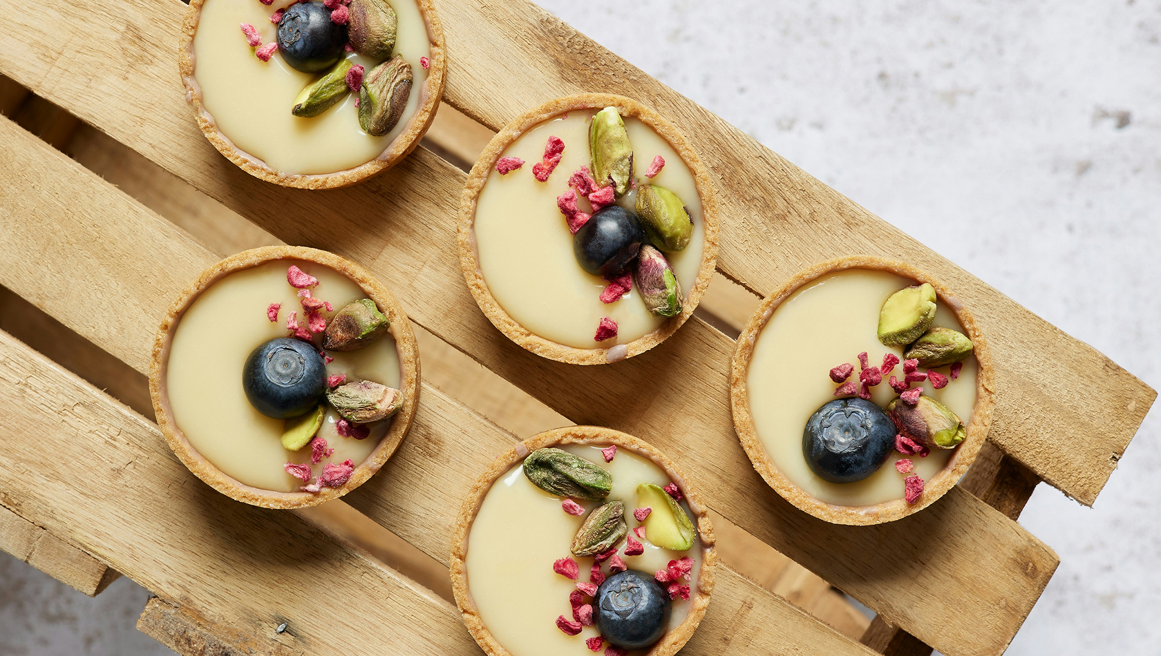 Fünf kleine Törtchen auf einem Holzbrett. Jedes Törtchen hat eine helle Füllung und ist mit Pistazien, blauen Beeren und gefriergetrockneten Himbeeren dekoriert.