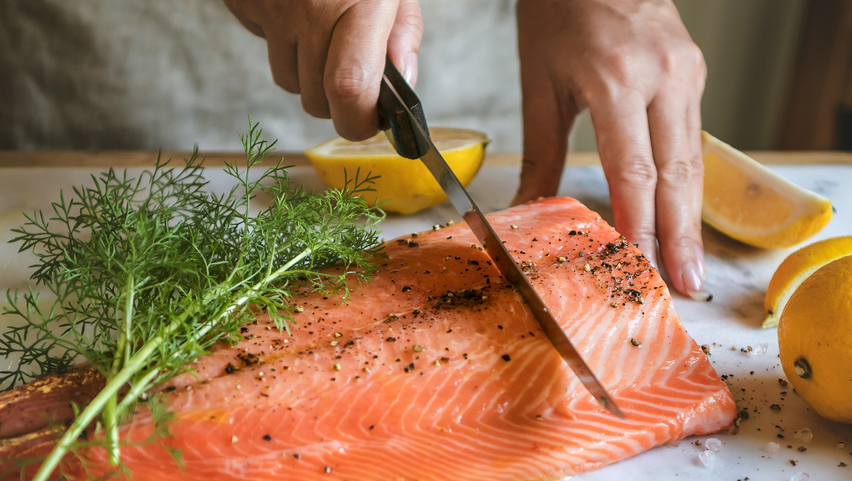 Eine Hand schneidet mit einem Messer einen Lachsfilet auf einem Tisch. Neben dem Fisch liegen frische Zitronen und etwas Dill. Der Lachs ist mit Pfeffer gewürzt.