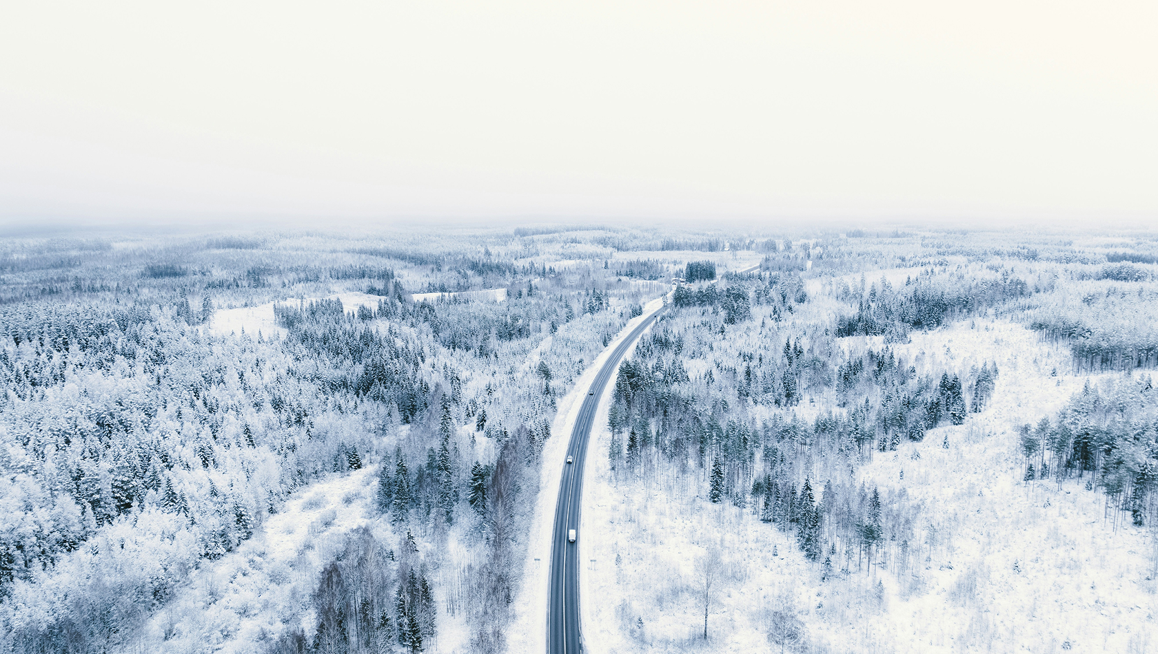 Luftaufnahme einer verschneiten Landschaft mit einem kurvigen Straßenverlauf. Umgeben von schneebedeckten Wäldern erstreckt sich die Straße in die Ferne, während der Himmel grau und neblig ist. Die gesamte Szene vermittelt eine ruhige, winterliche Atmosphäre.