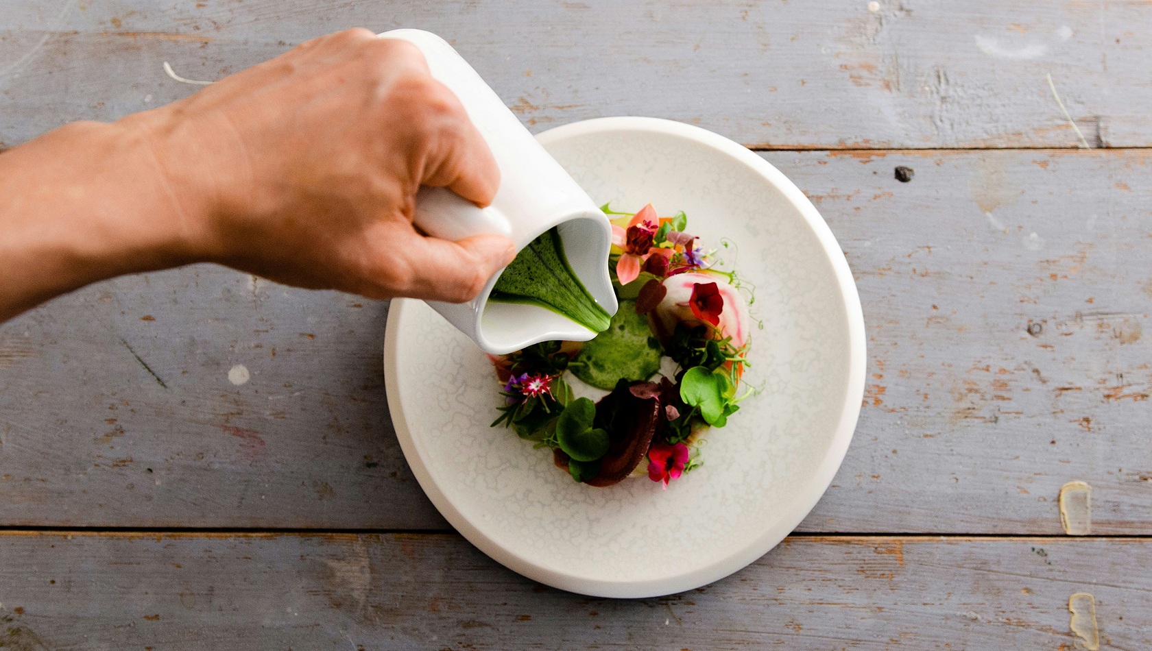 Eine Hand gießt eine grüne Sauce aus einem weißen Gefäß über einen schönen Teller mit frischen Kräutern und essbaren Blumen. Der Teller ist hell und rund, und die Zutaten sind kunstvoll angeordnet, was eine appetitliche Präsentation zeigt.