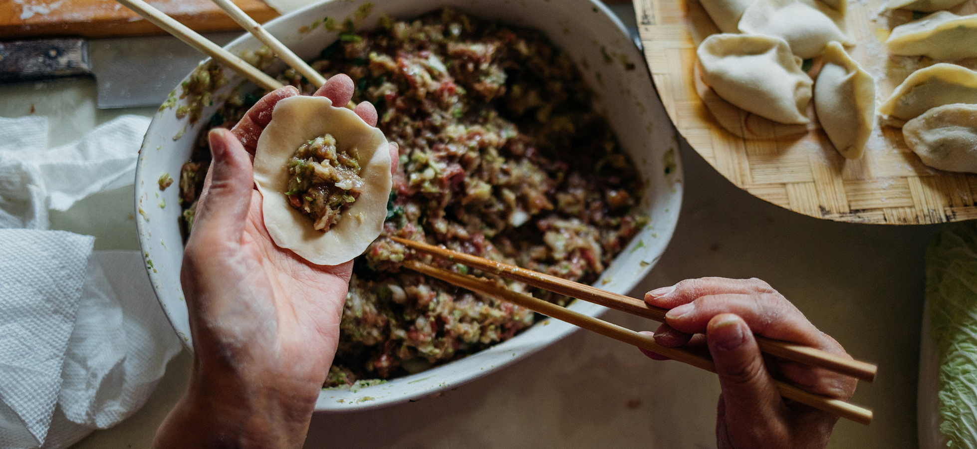 Eine Hand hält einen Teigkreis mit Füllung, während die andere Hand Essstäbchen benutzt. Im Hintergrund ist eine Schüssel mit einer Mischung aus gemahlenem Fleisch und Gemüse zu sehen, sowie ungeformte Teigtaschen auf einem Teller.