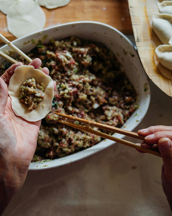 Eine Hand hält einen Teigkreis mit einer Füllung aus Fleisch und Gemüse, während die andere Hand Essstäbchen benutzt. Im Hintergrund befindet sich eine Schüssel mit der Füllmasse und bereits vorbereitete Teigtaschen.