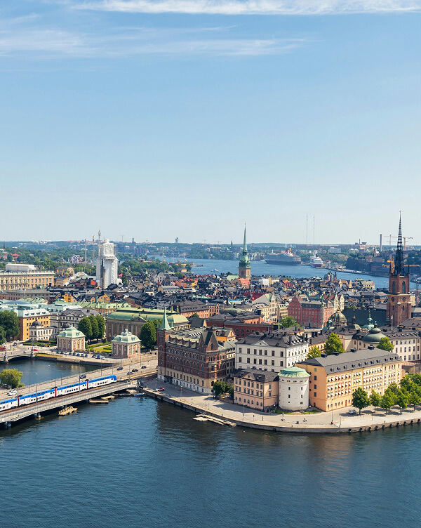 Eine Panoramasicht auf Stockholm, die Gebäude und Sehenswürdigkeiten der Altstadt zeigt. Im Vordergrund verläuft eine Brücke über das Wasser, während im Hintergrund die Schiffe im Hafen und grüne Landschaften zu sehen sind. Der Himmel ist klar und blau.