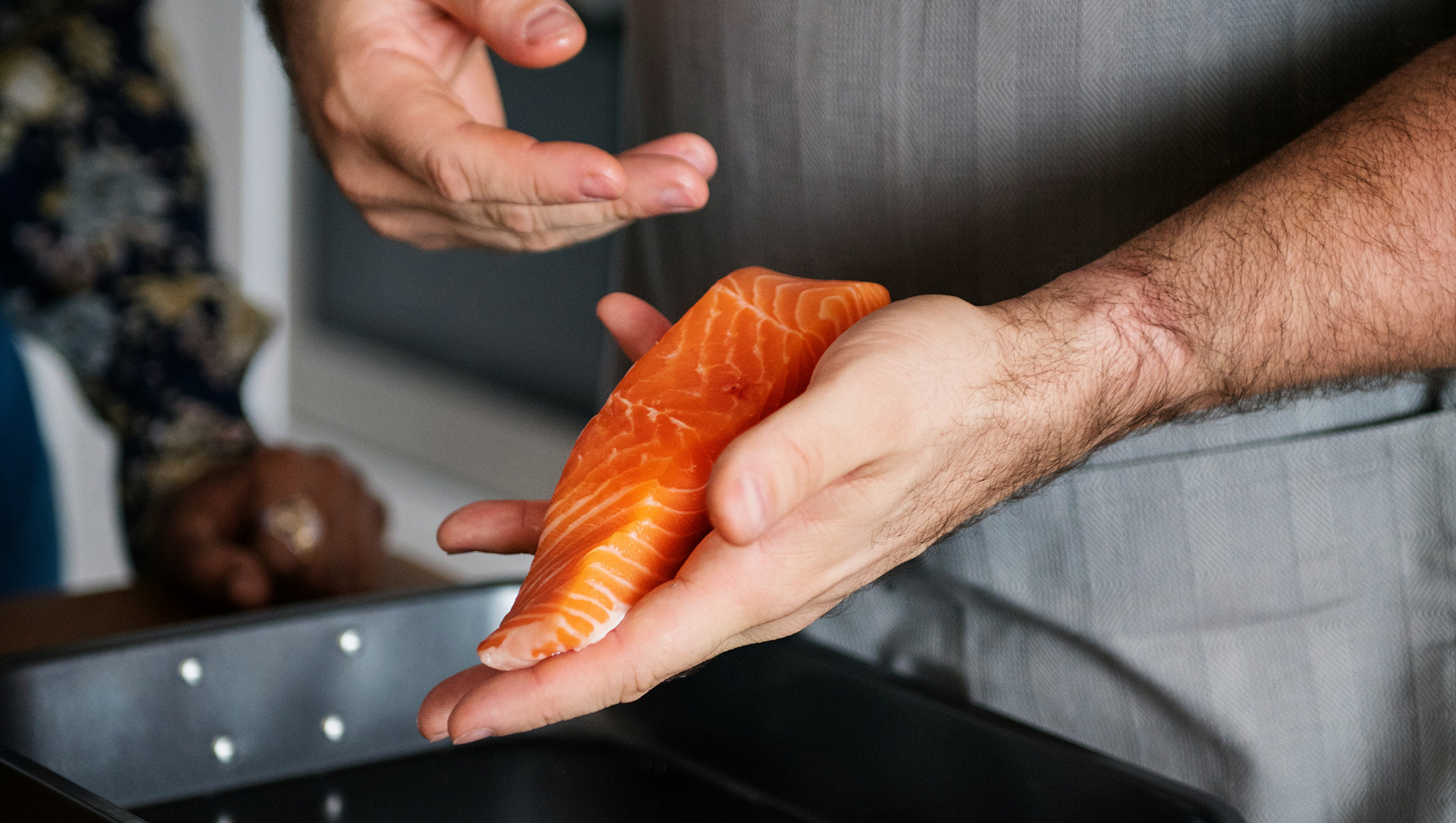 Eine Person hält ein frisches Stück Lachs in der Hand und zeigt es, während sie mit einer anderen Person spricht. Im Hintergrund ist ein Backblech sichtbar. Die Szene vermittelt einen Moment des Kochens oder der Zubereitung von Lebensmitteln.