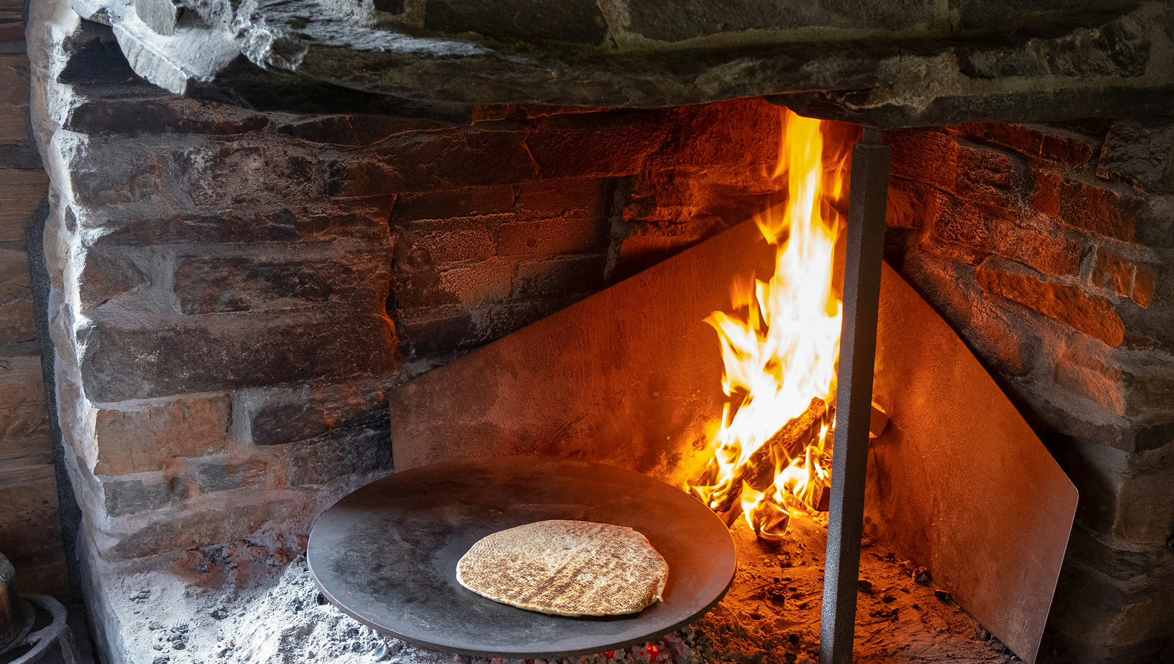 Ein flacher Teigfladen liegt auf einer metallenen Platte vor einem offenen Feuer in einem gemauerten Kamin. Die Flammen lodern, während der Teig gebacken wird. Die Szene vermittelt eine traditionelle, rustikale Kochumgebung.