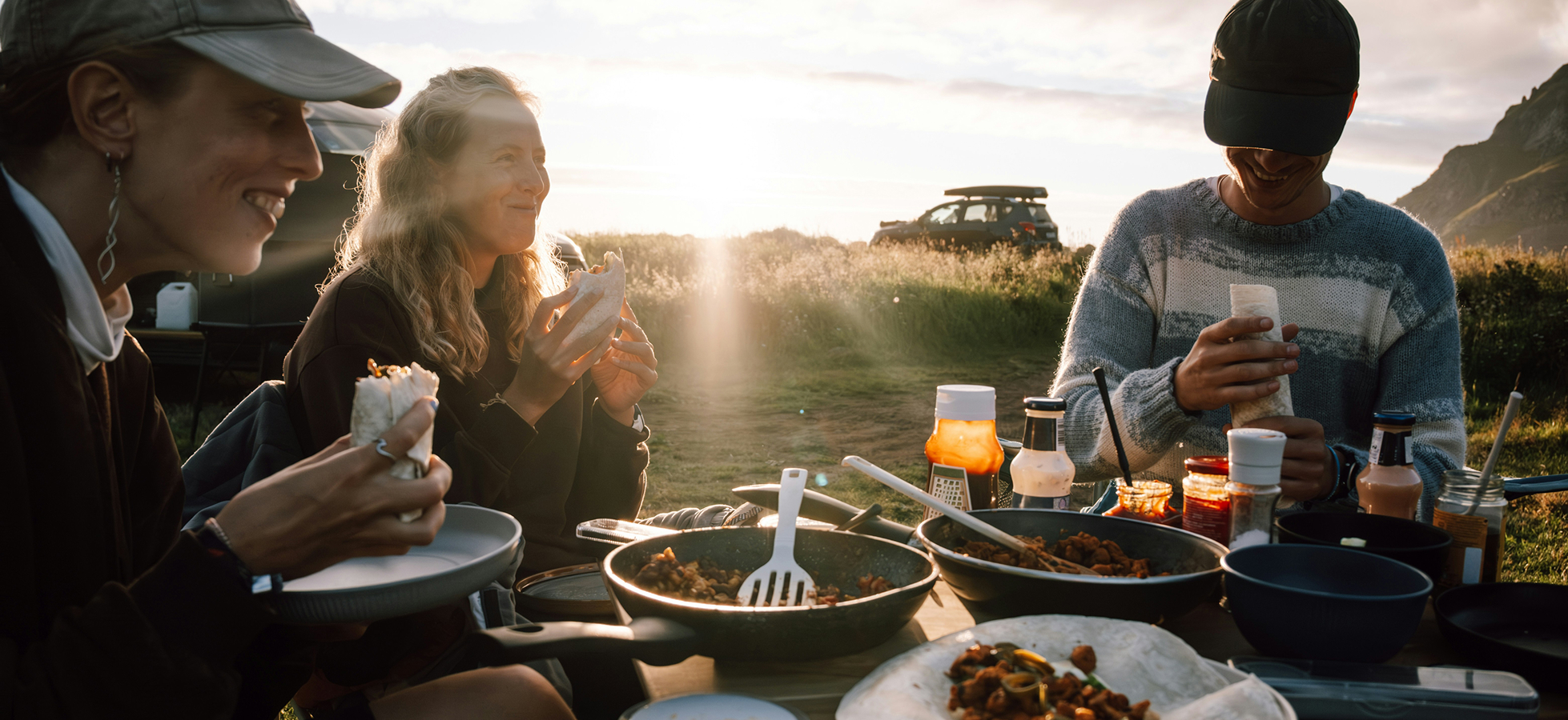 Drei Menschen sitzen am Tisch im Freien und genießen eine Mahlzeit. Sie halten verschiedene Speisen in den Händen, während die Sonne im Hintergrund aufgeht. Vor ihnen stehen Töpfe und Flaschen mit Lebensmitteln. Die Szenerie vermittelt ein Gefühl der Geselligkeit und Naturverbundenheit.