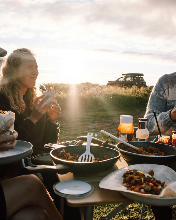 Eine Gruppe von Personen sitzt im Freien und genießt eine Mahlzeit. Auf einem Tisch stehen verschiedene Töpfe mit Essen und Saucen. Die Sonne scheint im Hintergrund und schafft eine warme Atmosphäre. Alle scheinen miteinander zu kommunizieren und die Zeit zu genießen.