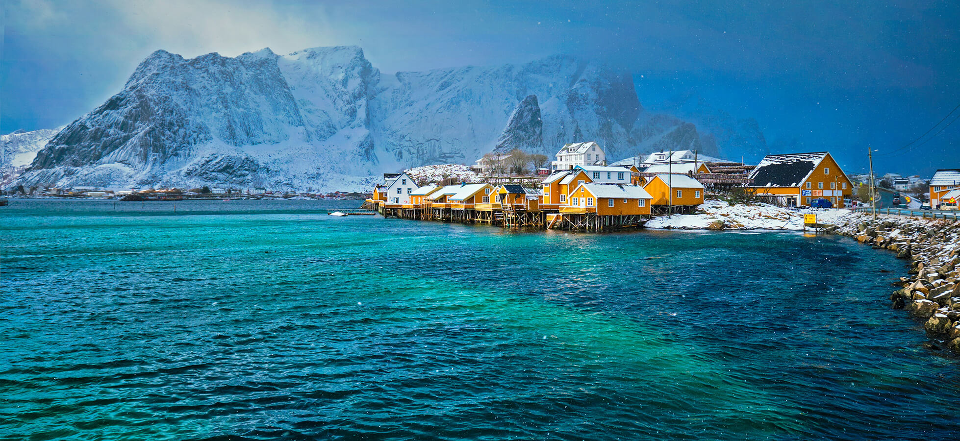 Eine malerische Küstenlandschaft mit gelben Holzhäusern am Wasser, umgeben von schneebedeckten Bergen. Die ruhige, grünlich-blaue See reflektiert die winterliche Atmosphäre unter einem bewölkten Himmel.
