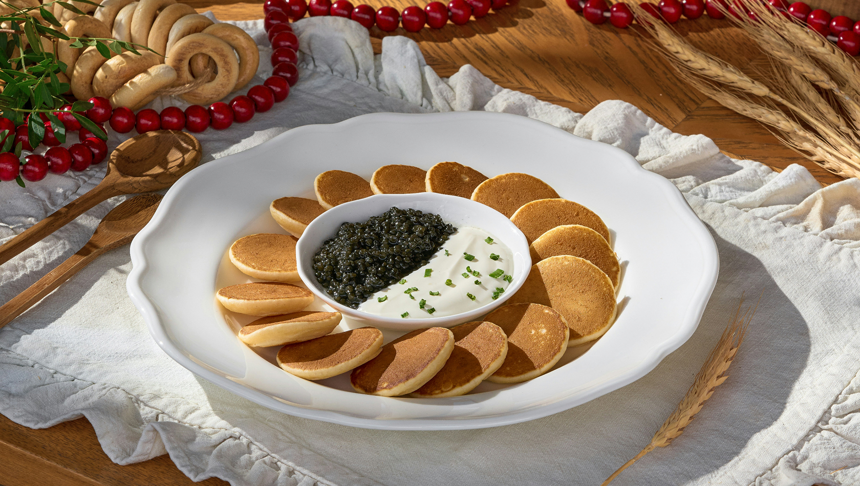 Ein weißer Teller mit kleinen Stücken von gefalteten Pfannkuchen um einen zentralen Dip aus Sauerrahm und Kaviar. Dekorative Elemente wie Brot und rote Beeren sind im Hintergrund sichtbar. Die Anordnung vermittelt ein elegantes und einladendes Ambiente.
