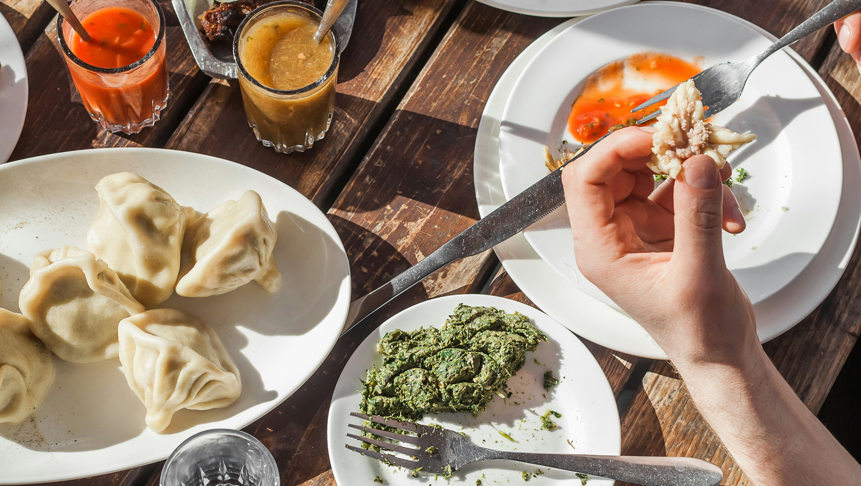 Ein Tisch mit verschiedenen Gerichten: Eine Hand hält ein Stück gefüllten Teig (Dumpling) und eine Gabel. Auf dem Tisch befinden sich auch zwei Saucen, ein Tellerrand mit grünem Pesto und ein weiterer Teller mit einer roten Sauce, alles auf einer Holzoberfläche.