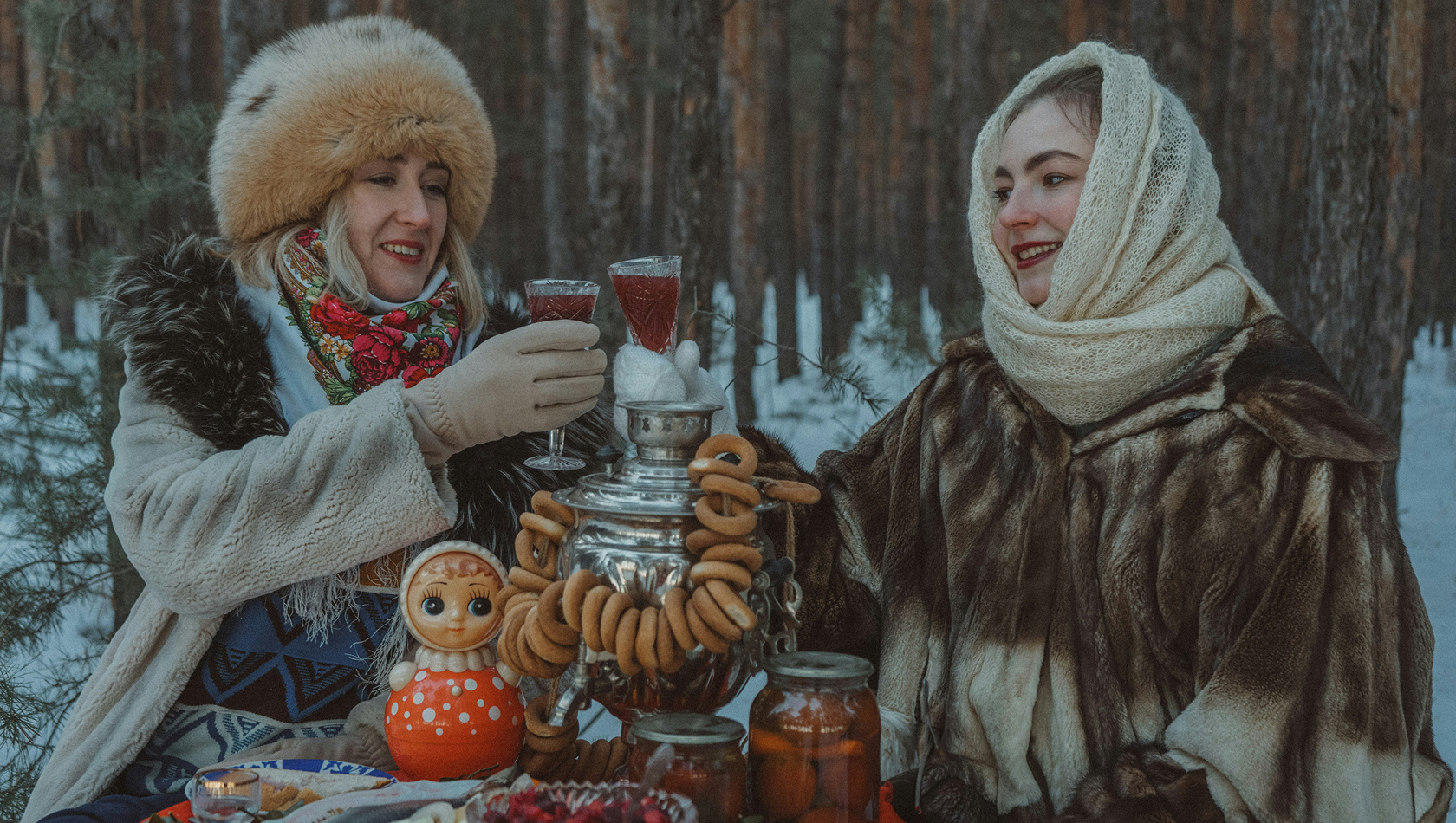 Zwei Frauen sitzen in einer winterlichen Waldlandschaft und stoßen mit Gläsern an. Sie tragen warme, traditionelle Kleidung. Vor ihnen steht eine Teekanne und verschiedene Speisen, darunter eine dekorative Matroschka-Puppe und Kekse. Die Szene strahlt Gemütlichkeit und Freundschaft aus.