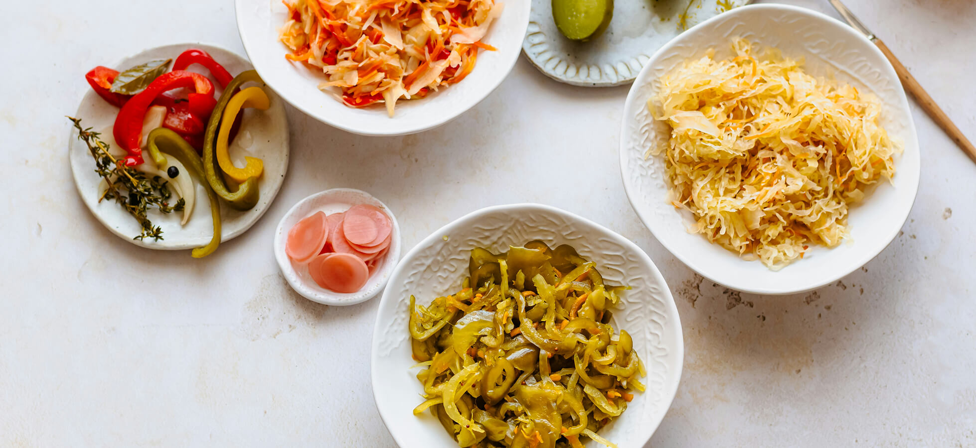Eine Auswahl an verschiedenen eingelegten Gemüsegerichten in Schalen. Dazu gehören gelbe und grüne Paprika, Sauerkraut und eine Art von eingelegten Zwiebeln. Dekoriert mit frischen Kräutern auf einem hellen Tisch.