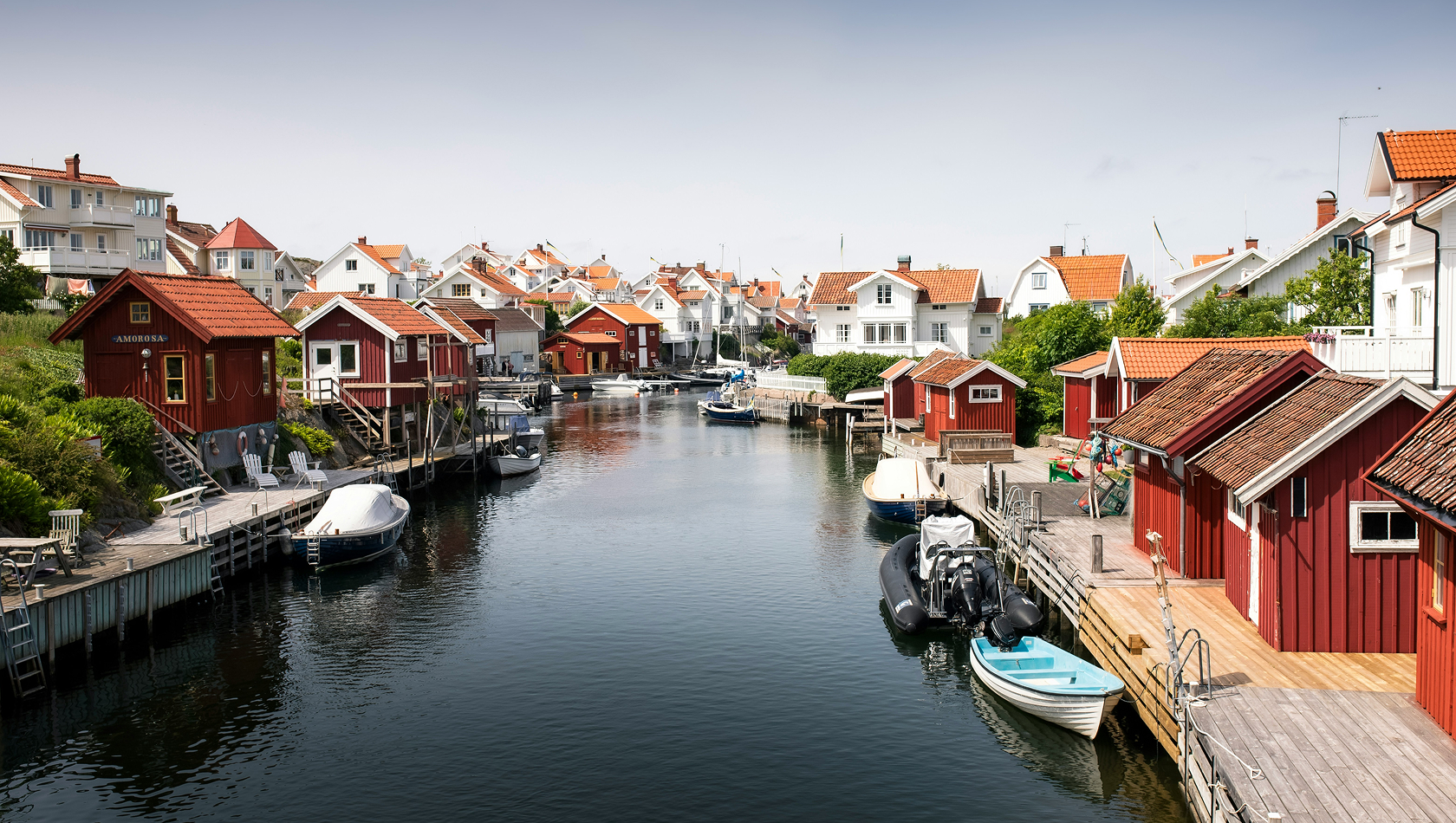 Ein malerischer Kanal mit roten Holzhäusern auf beiden Seiten. Boote liegen am Steg. Im Hintergrund sind weitere Häuser mit roten Dächern sichtbar. Die Szenerie vermittelt ein ruhiges, maritimes Ambiente.