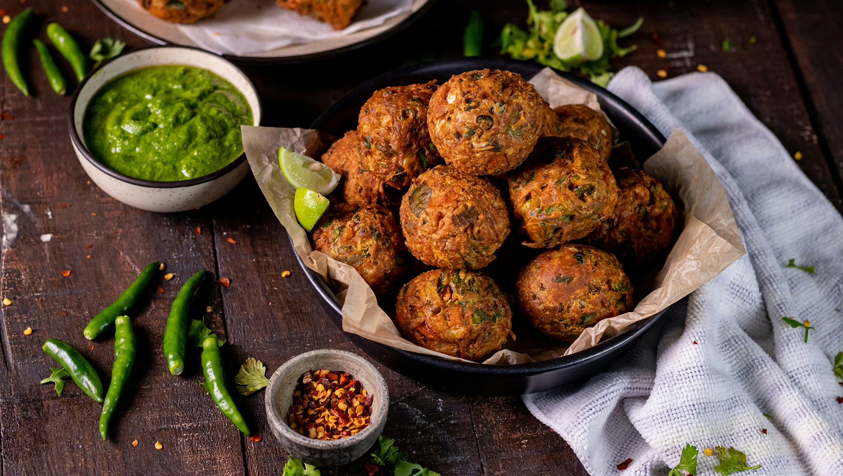 Eine schwarze Schüssel enthält mehrere frittierte Bällchen, die mit Kräutern und Gewürzen zubereitet sind. Neben der Schüssel liegt eine kleine Schale mit grüner Soße und eine weitere mit roten Chili-Flocken. Grünes Gemüse und einige Chilischoten sind auf dem Tisch verteilt.