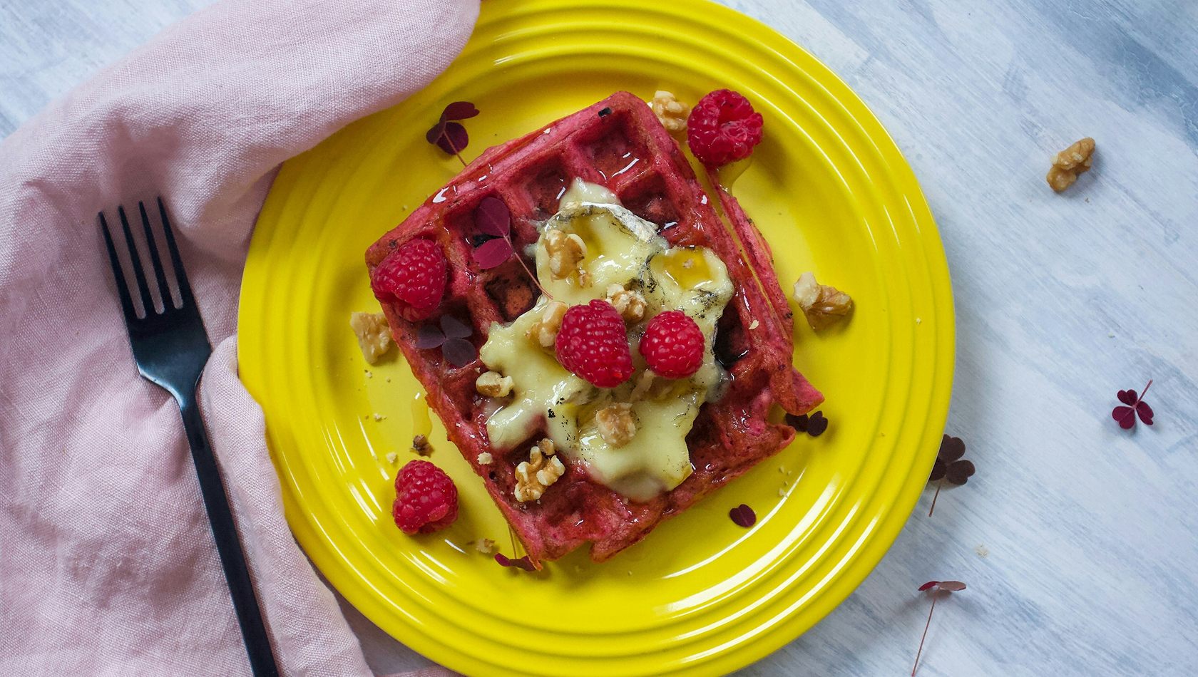 Ein Teller mit einer bunten Waffel, garniert mit frischen Himbeeren, gehackten Walnüssen und geschmolzenem Käse. Der Teller ist gelb und das Besteck, eine Gabel, liegt daneben. Im Hintergrund ist ein zarter Stoff in Pastellfarbe sichtbar.