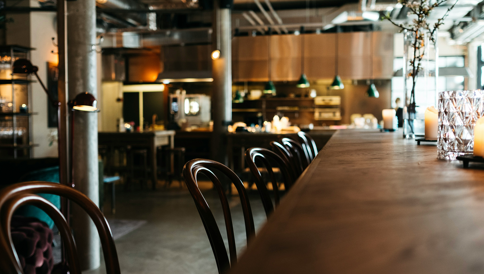 Ein gemütliches Café mit einer langen Holzbar, an der mehrere Stühle stehen. Im Hintergrund sind ein offenes Küchenbereich und warme Lichtquellen zu sehen. Die Atmosphäre ist einladend und modern, ideal für gesellige Treffen oder entspannte Stunden.