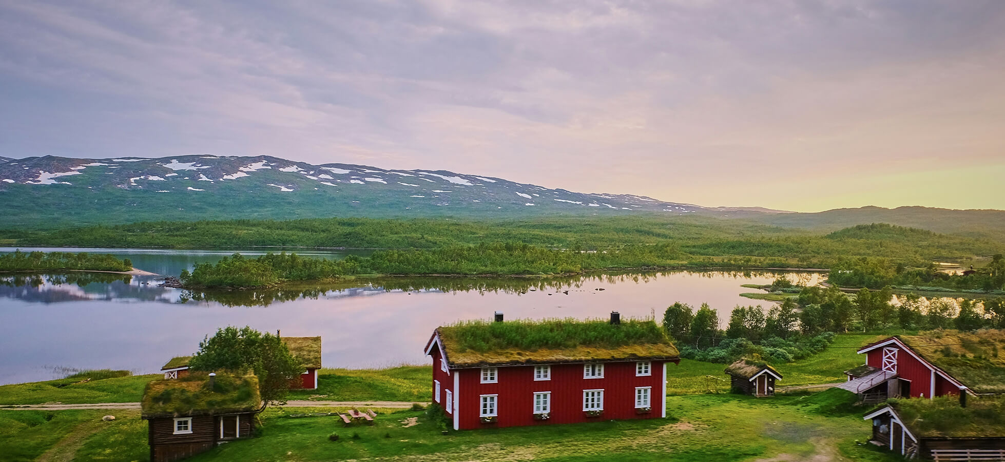 Eine ländliche Landschaft mit roten, strohgedeckten Häusern am Ufer eines ruhigen Sees. Im Hintergrund erstrecken sich grüne Hügel und schneebedeckte Berge, während der Himmel in sanften Pastelltönen leuchtet. Die Szene strahlt eine ruhige, naturnahe Atmosphäre aus.