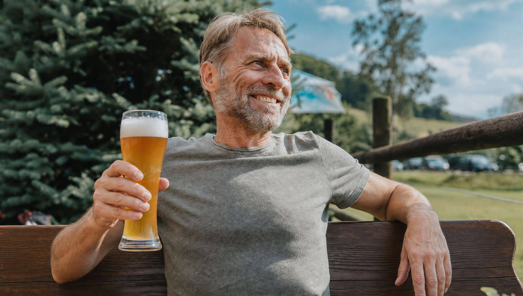 Mann mittleren Alters sitzt entspannt auf einer Bank im Freien, lächelt und hält ein hohes Bierglas in der Hand.