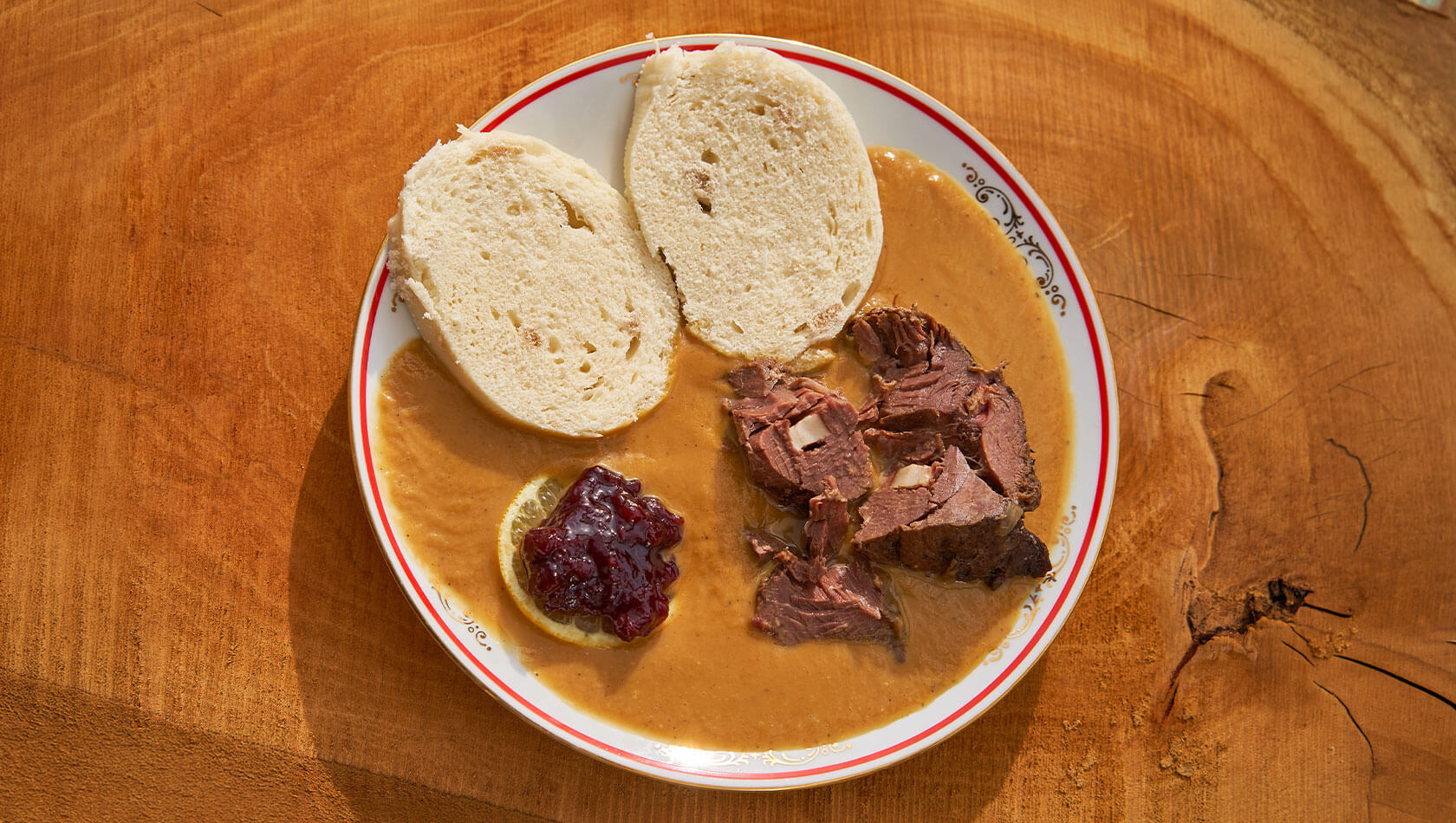 Teller mit drei hellen Knödel- oder Pfannkuchenstücken, übergossen von cremiger gelber Sauce; in der Mitte kleines rundes Gebäck mit roten Beeren und Sahne, daneben ein Stück dunkler Kuchen oder Brot.
