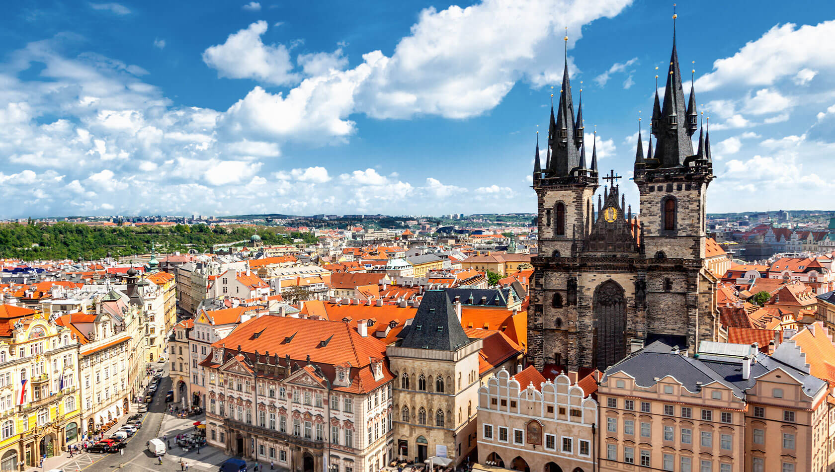 Panoramablick auf die Prager Altstadt: markante gotische Teynkirche mit zwei spitzen Türmen über roten Dachlandschaften und historischen Fassaden, unter blauem Himmel mit weißen Wolken.