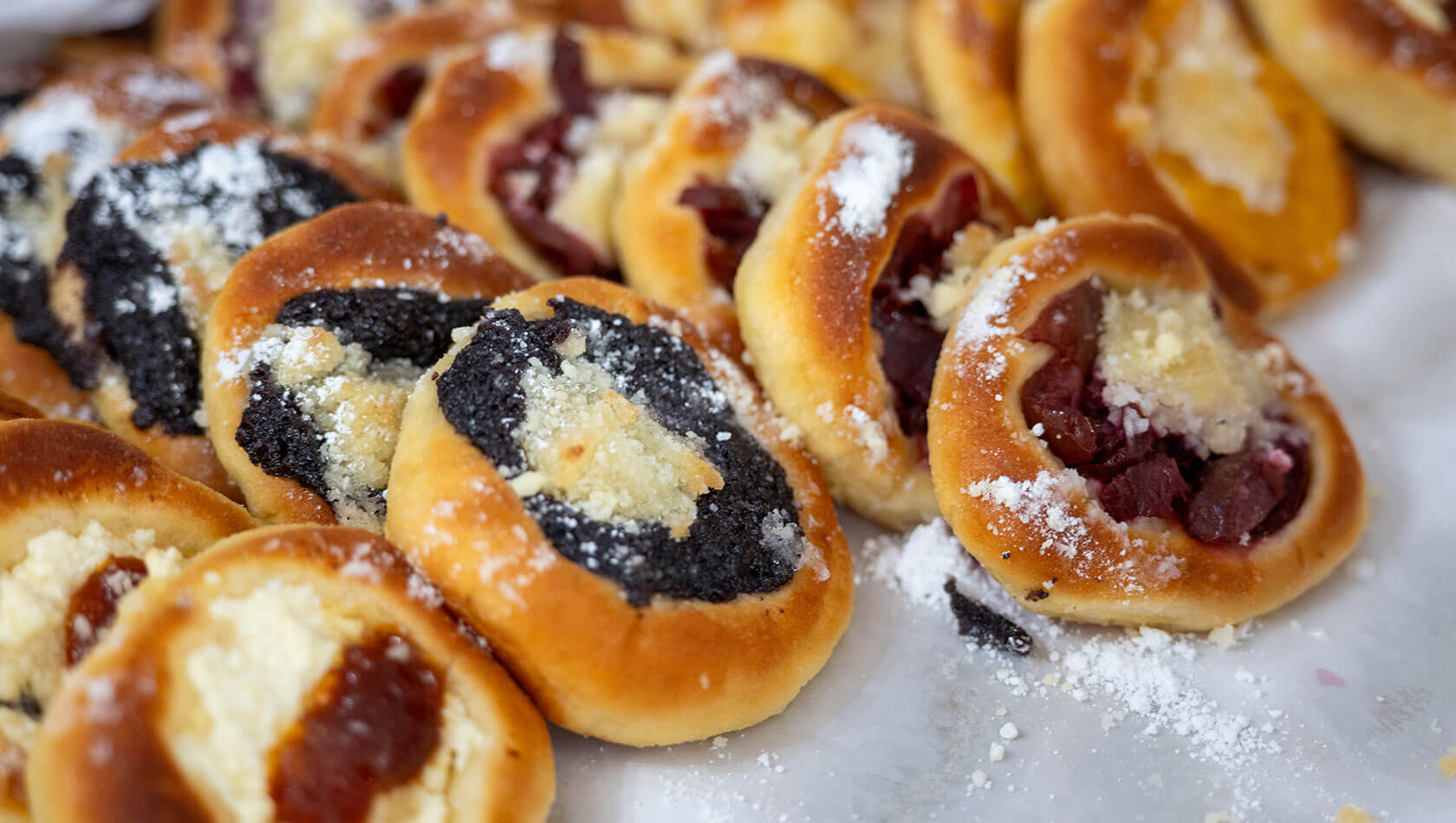 Auflaufform mit goldbraunen, zart zuckerbestäubten Hefebrötchen (Buchteln); daneben ein Glas Marmelade mit Löffel.