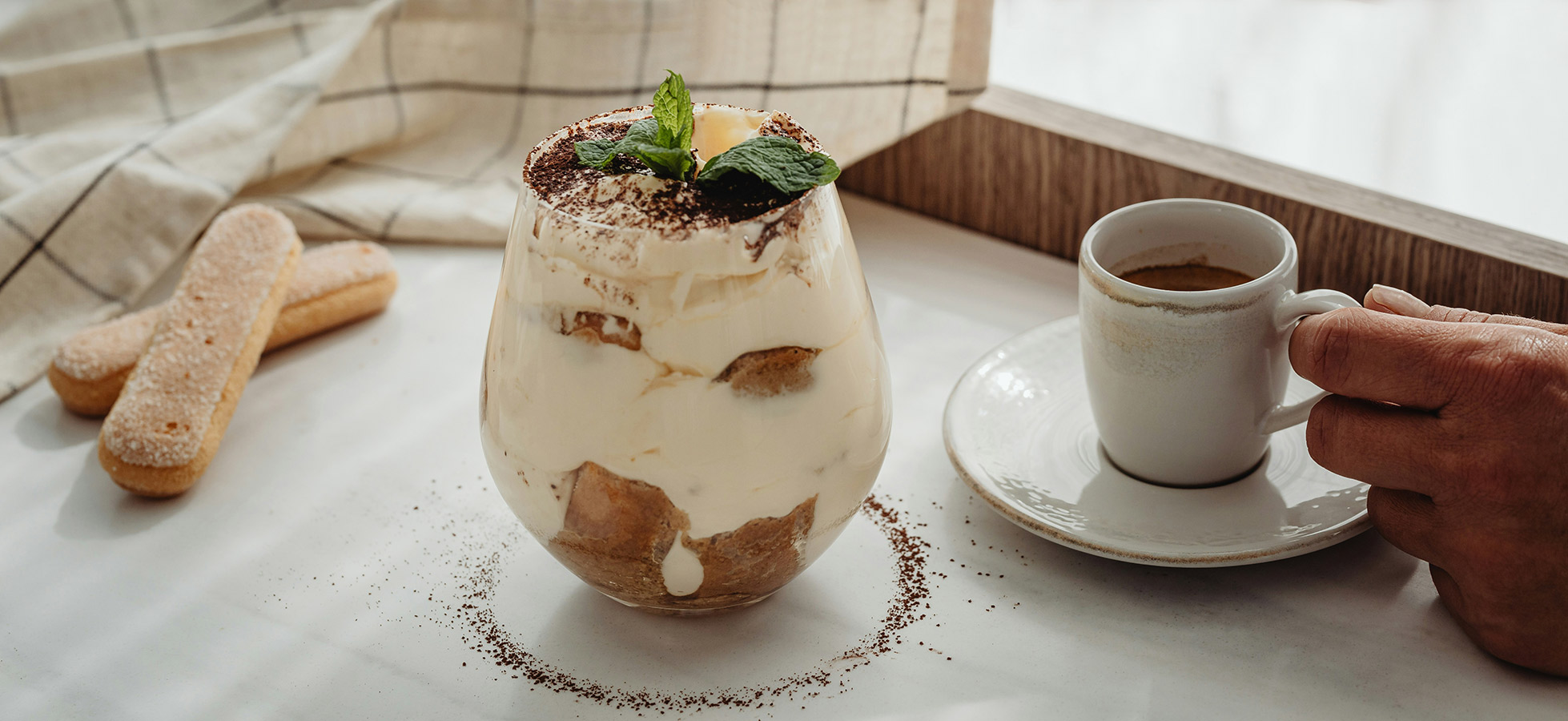 Ein Glas mit einer Schichtdessert aus Sahne, Keksen und Kakaopulver, garniert mit frischen Minzblättern. Eine Hand hält einen Löffel, um das Dessert zu servieren. Neben dem Glas steht eine Tasse Kaffee. Im Hintergrund ist ein karierter Stoff sichtbar.