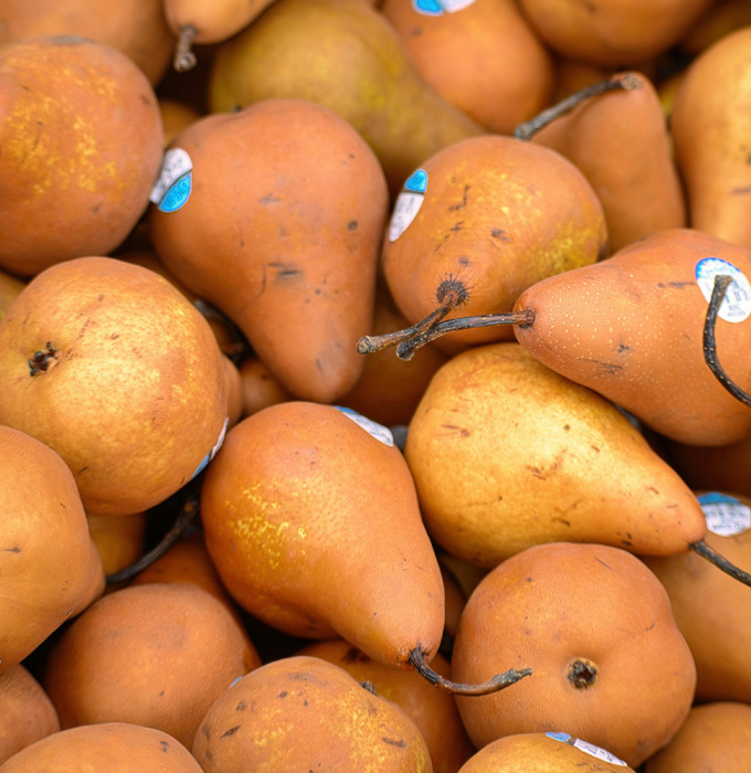 Eine Vielzahl von reifen, orangenen Birnen liegt übereinander. Einige tragen blaue Etiketten. Die Birnen haben eine glatte Haut und sind in verschiedenen Positionen angeordnet, was eine kräftige und frische Präsentation ergibt.