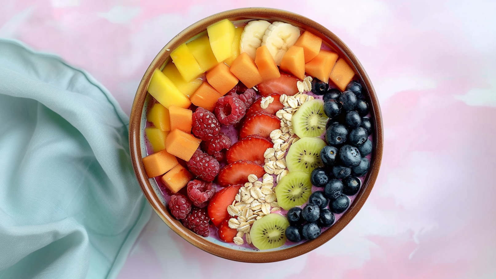 Eine Schüssel mit einer bunten Mischung aus frischen Früchten: Mango, Banane, Melone, Himbeeren, Erdbeeren, Kiwi und Heidelbeeren. Darüber liegen Haferflocken. Der Hintergrund ist sanft rosa.