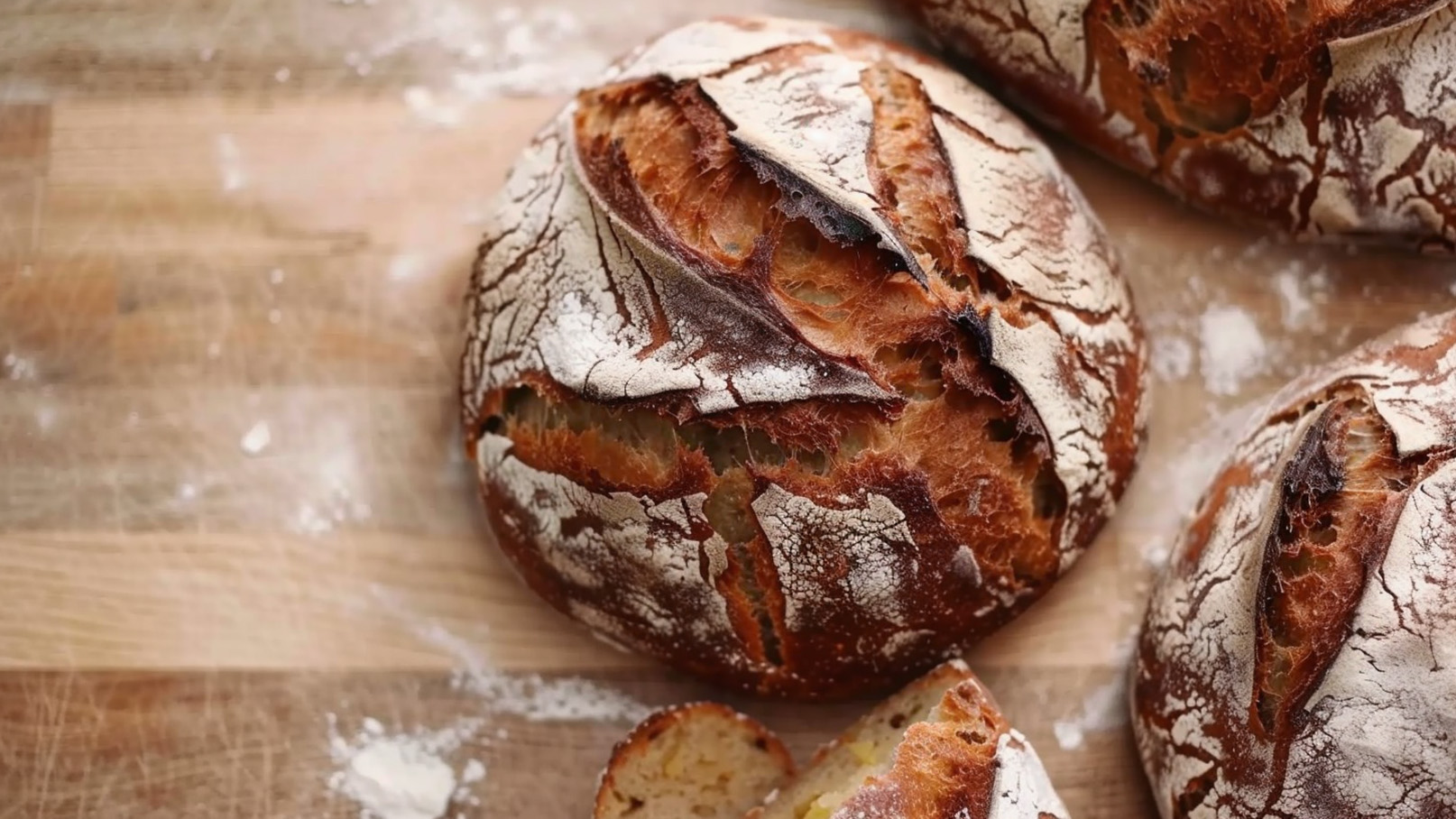 Auf dem Bild sind mehrere runde, frisch gebackene Brote zu sehen. Die Brote haben eine rustikale Kruste mit Rissen und einer leichten Mehlschicht. Ein Brot ist aufgeschnitten, um die weiche, goldene Innenseite zu zeigen. Der Hintergrund ist eine Holzoberfläche, die mit etwas Mehl bestäubt ist.