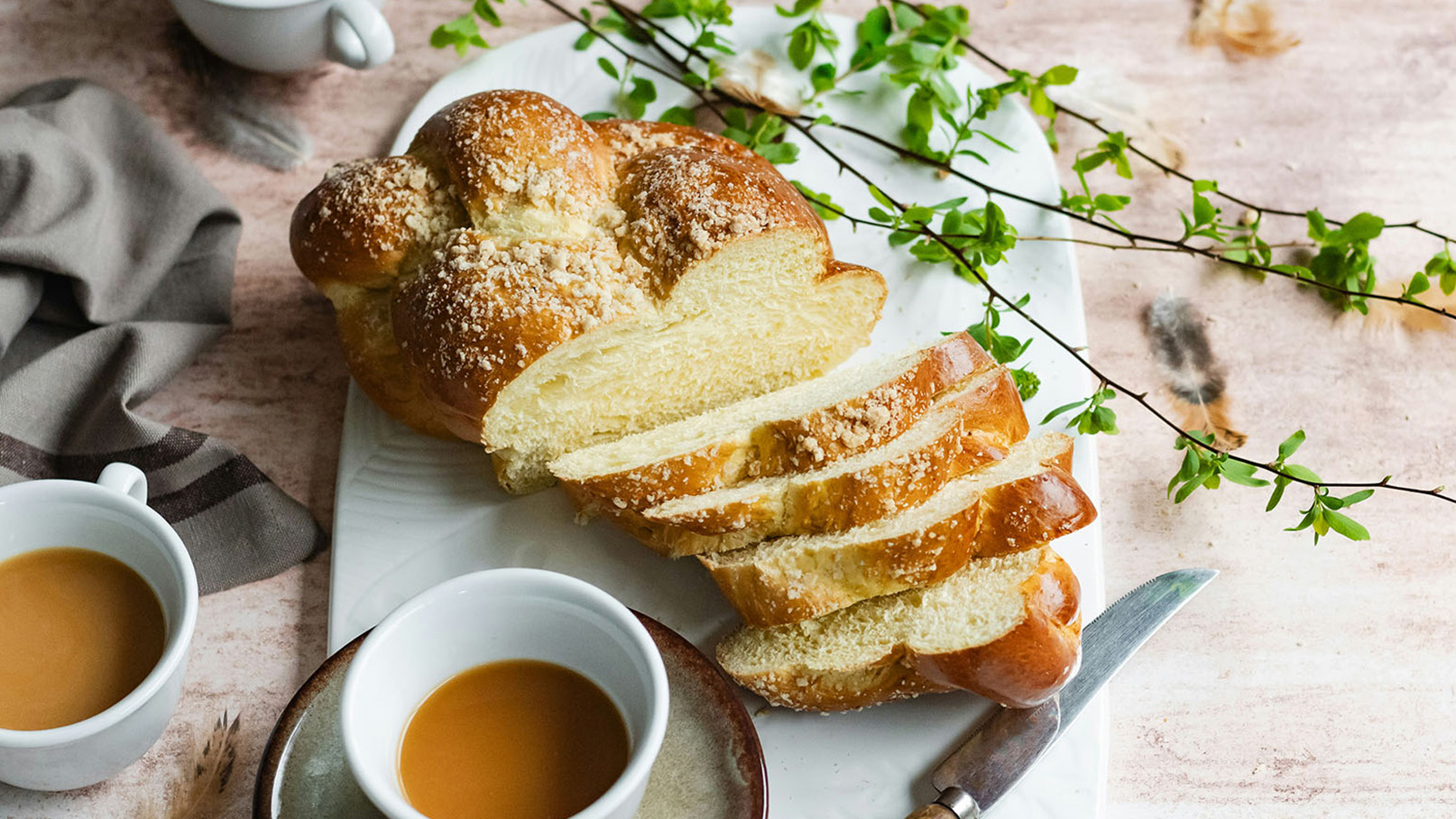 Ein frisch gebackener, geflochtener Zopf aus hellem Brot liegt auf einem weißen Teller. Ein Teil des Zopfs ist in Scheiben geschnitten. Daneben stehen zwei Tassen mit Kaffee auf einem Holzuntergrund, begleitet von etwas frischem Grün.