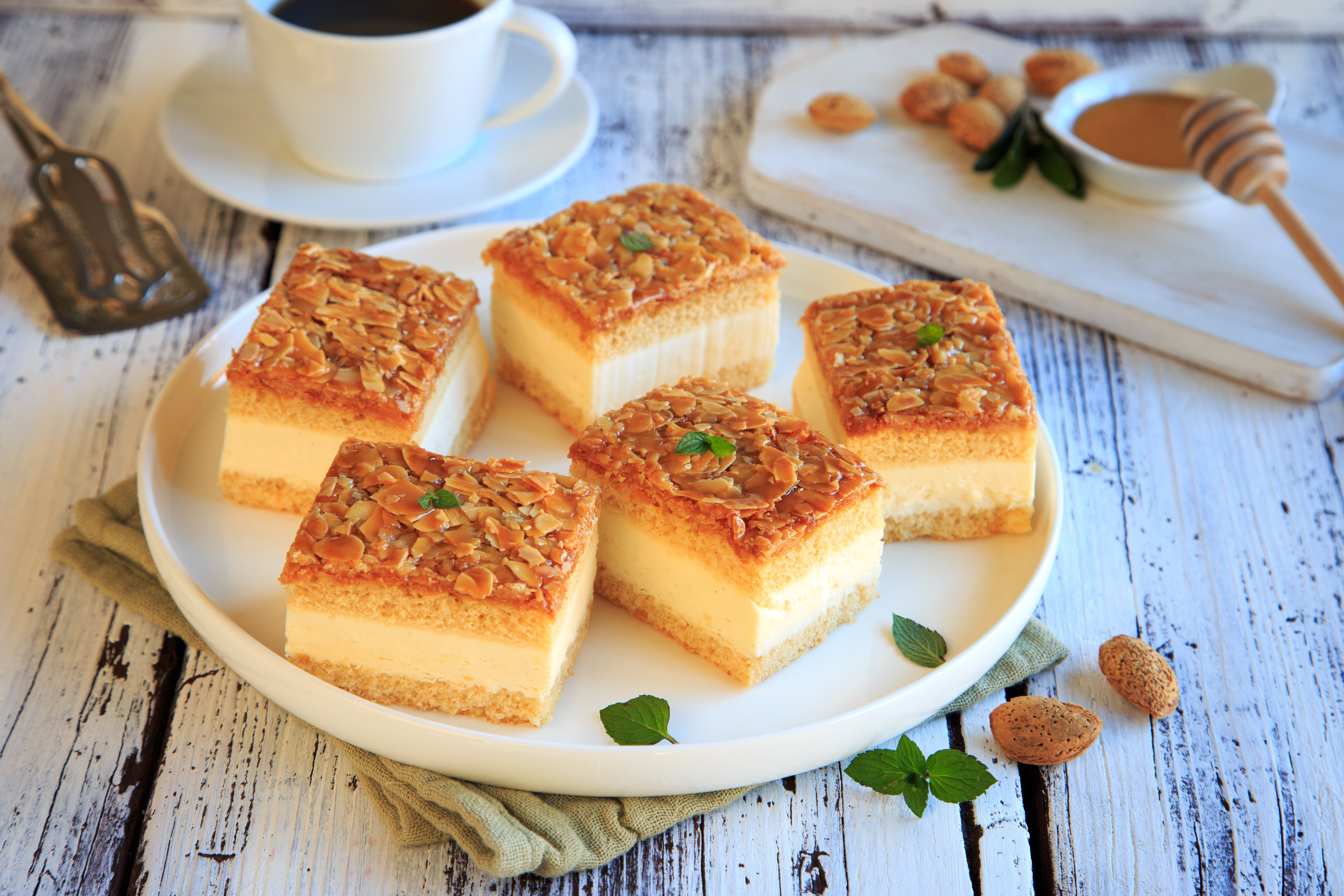Auf einem weißen Teller liegen fünf Stücke einer Dessert-Spezialität, die mit karamellisierten Mandeln und einer cremigen Füllung geschichtet sind. Frische Minzblätter dekorieren die Stücke. Im Hintergrund steht eine Tasse Kaffee und eine kleine Schale mit Honig und Mandeln.