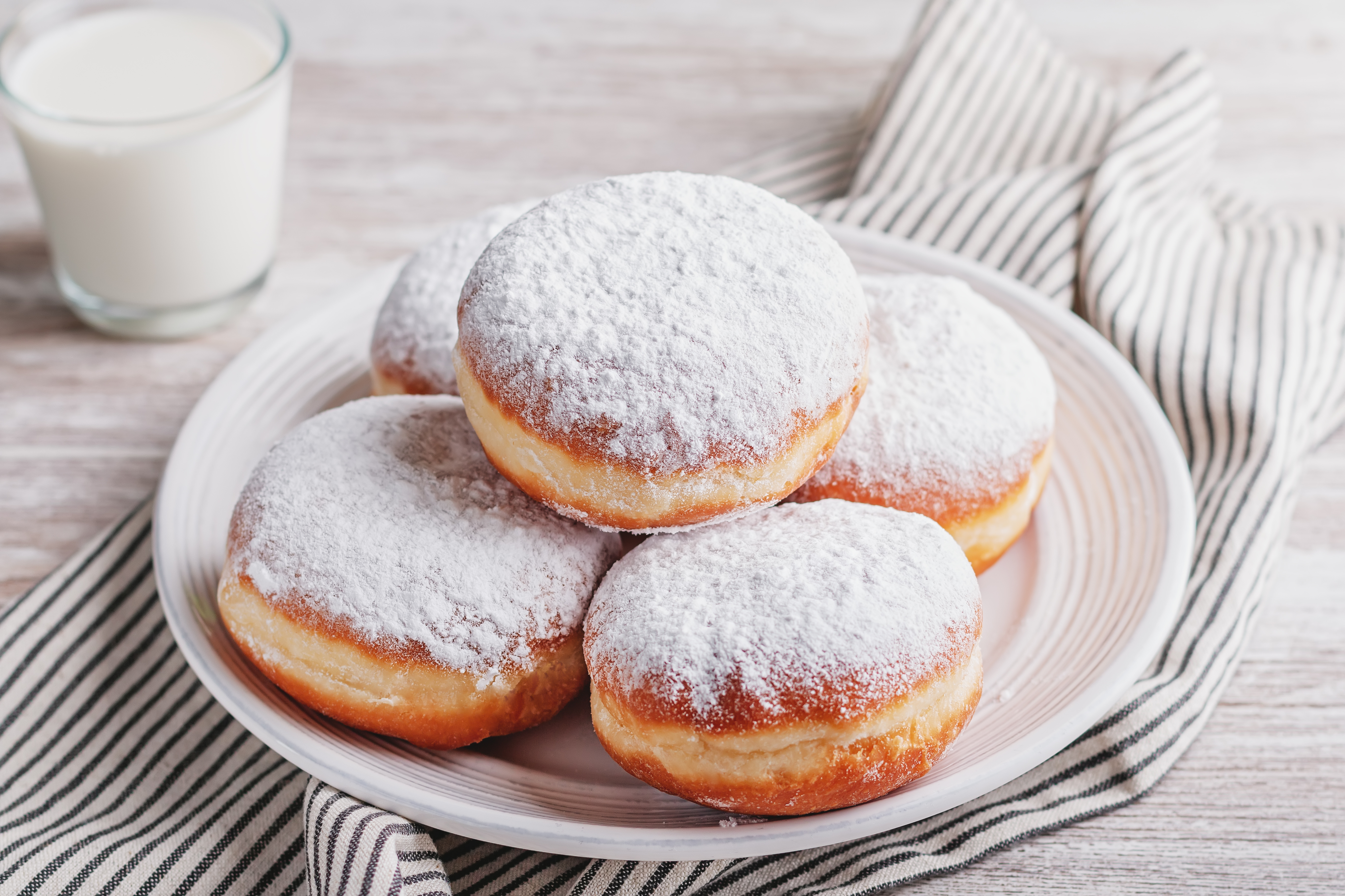 Auf dem Bild sind mehrere frisch gebackene Berliner zu sehen, die mit Puderzucker bestäubt sind. Sie liegen auf einem weißen Teller und sind rund und goldbraun. Im Hintergrund steht ein Glas Milch.