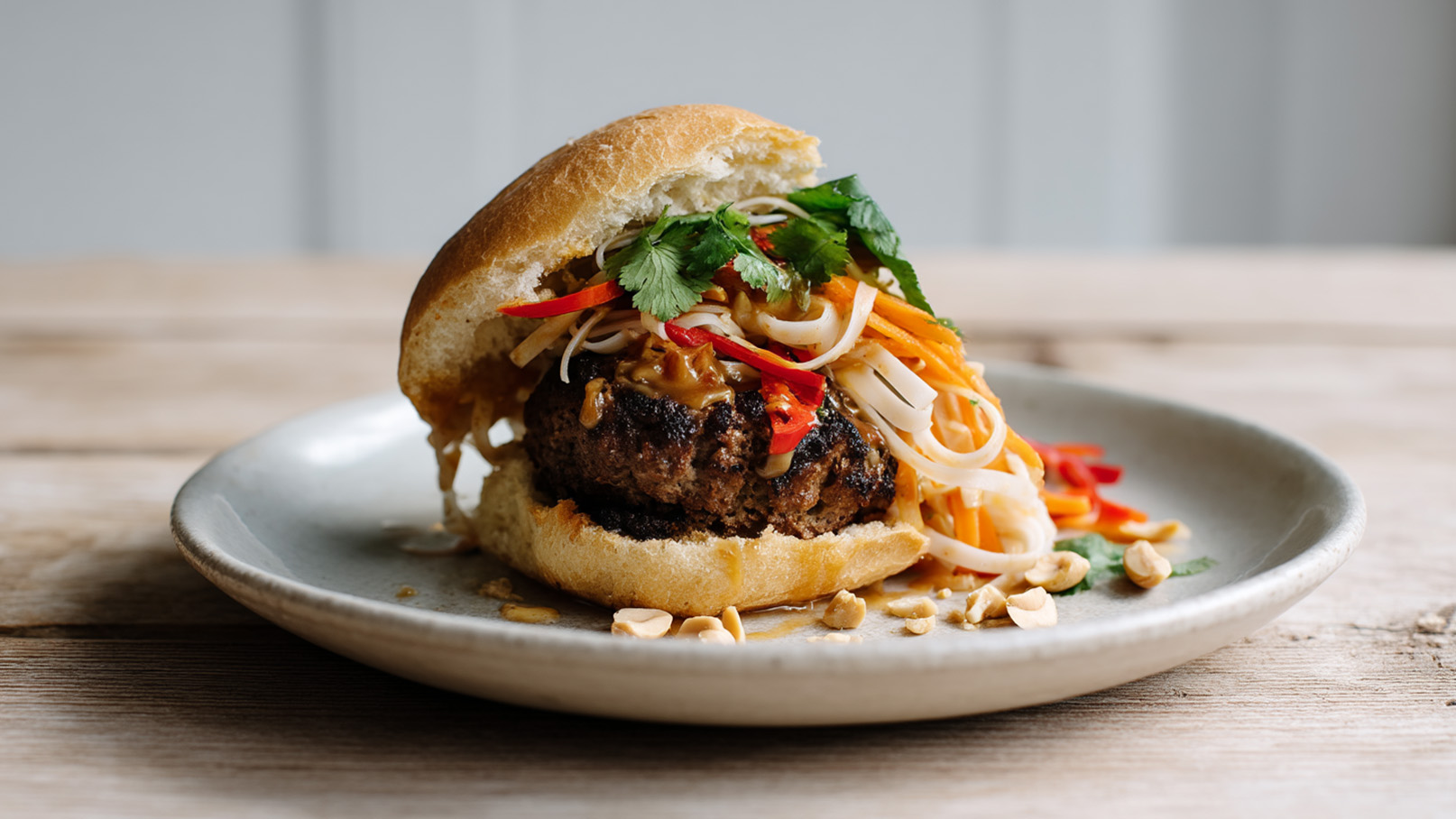 Ein frisch zubereiteter Burger auf einem Teller, mit einem saftigen Patty, frischem Gemüse, reifen Kräutern und einer bunten Mischung aus Nudeln und Saucen. Der Burger ist im Brötchen gefüllt und wird von gehackten Erdnüssen umgeben.