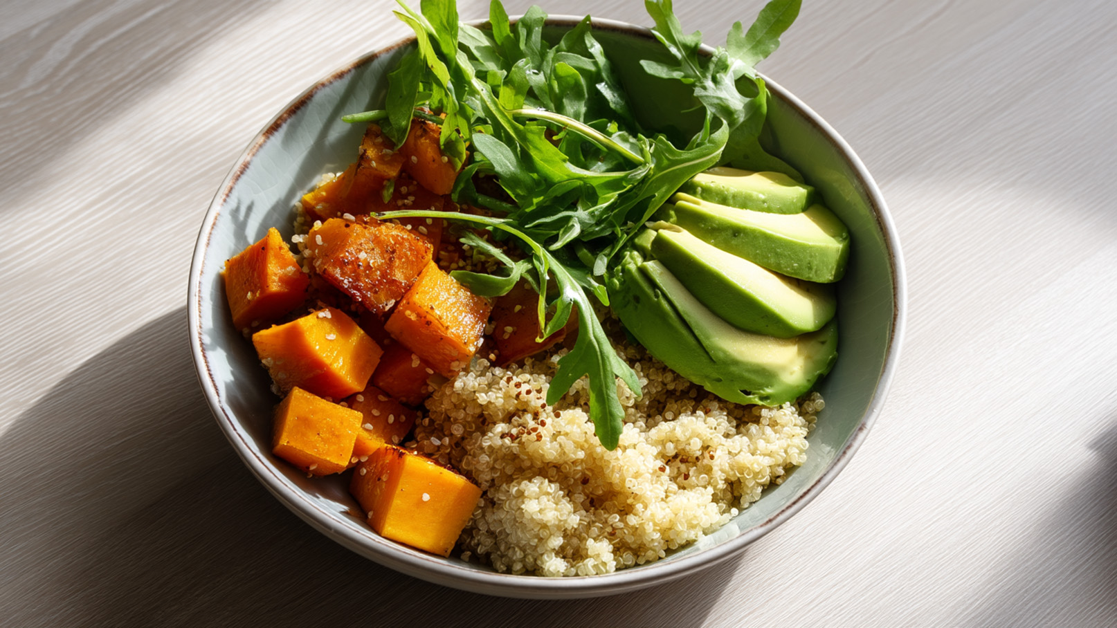 Eine Schüssel mit Quinoa, Kürbiswürfeln und Avocadoscheiben, garniert mit frischem Rucola. Die Zutaten sind bunt und gesund, auf einer hellen Tischoberfläche.