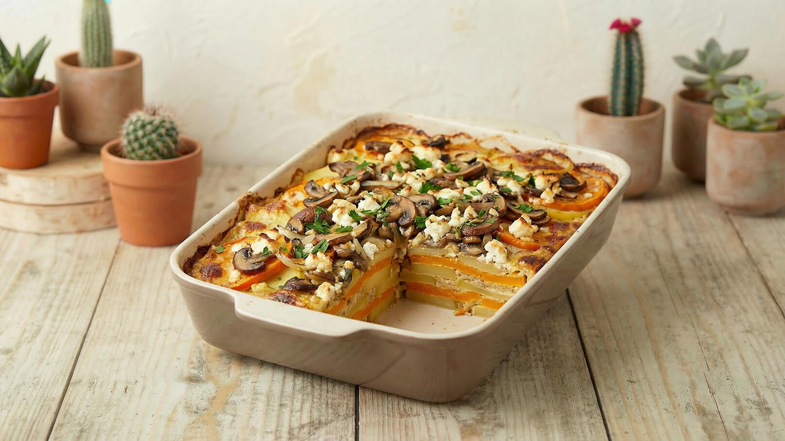 Ein Auflauf in einer beigefarbenen Form, schichtweise gefüllt mit Kürbis, Kartoffeln und Pilzen, überbacken mit Käse und frischen Kräutern. Der Auflauf ist bereits angeschnitten und auf einem Holzuntergrund platziert. Im Hintergrund sind einige Tontöpfe mit Pflanzen zu sehen.
