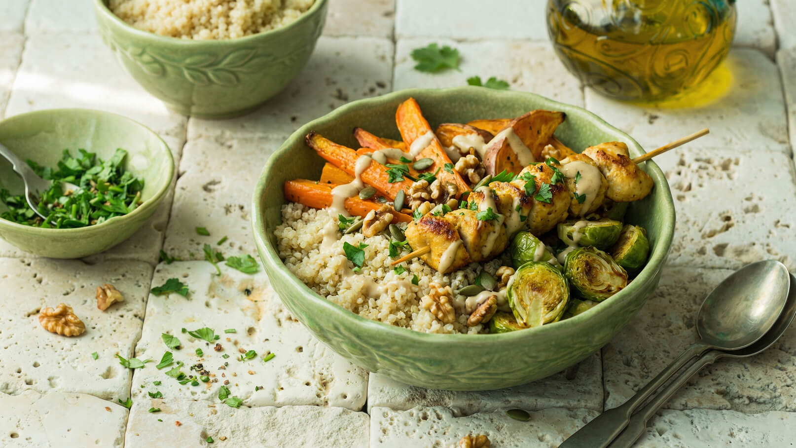 Eine grüne Schüssel enthält Quinoa, gegrillte Hähnchenspieße, geröstete Karotten, Rosenkohl und Walnüsse, garniert mit Kräutern. Daneben steht eine kleine Schüssel mit gehackten Kräutern und eine Gabel. Der Untergrund ist aus hellen Fliesen.