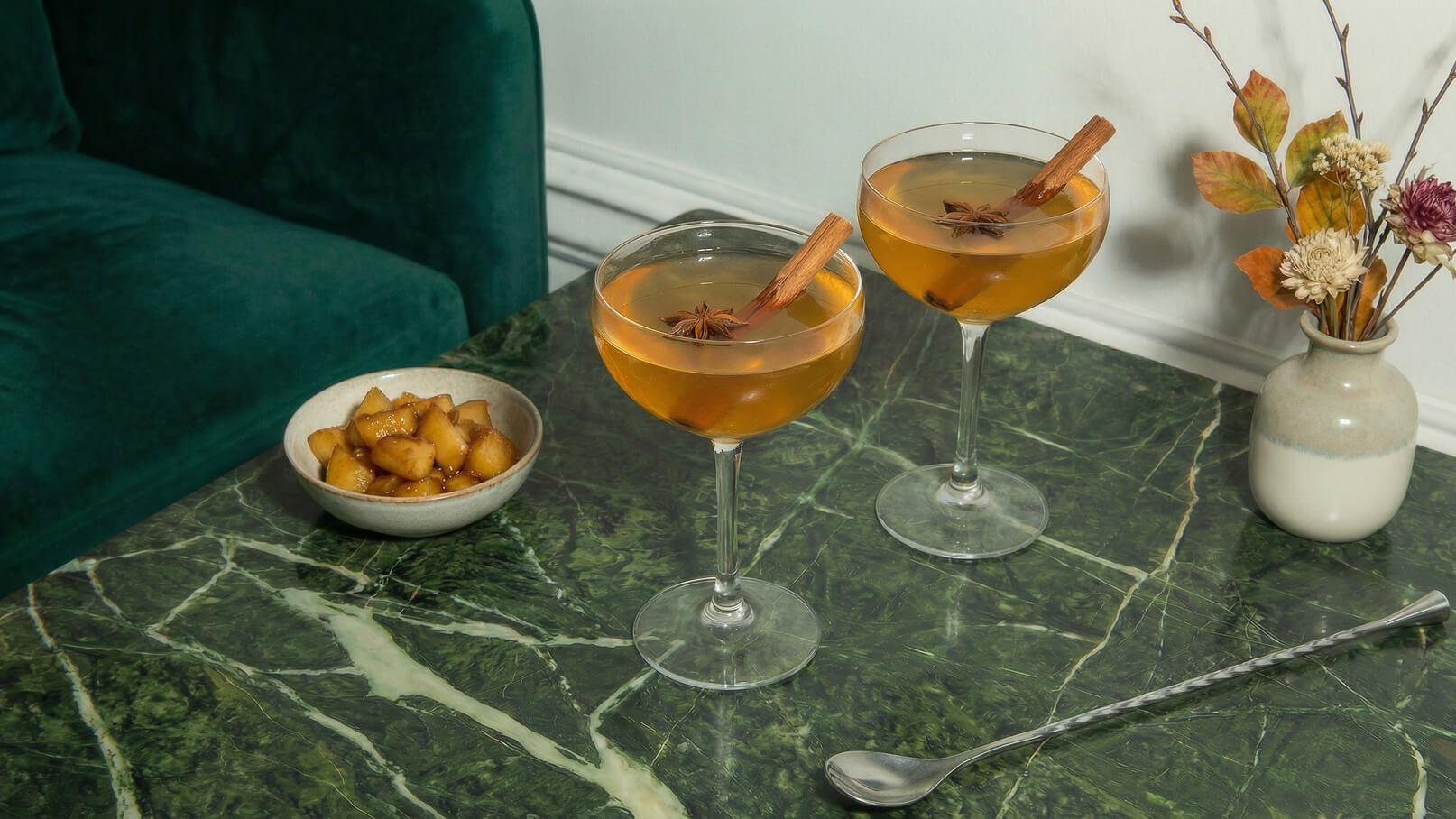 Zwei Cocktailgläser mit einem goldenen Getränk stehen auf einem marmorierten Tisch. In jedem Glas ist ein Zimtstängel und eine Sternanis zu sehen. Neben den Gläsern befindet sich eine kleine Schüssel mit karamellisierten Äpfeln. Im Hintergrund liegt eine grünen Couch.