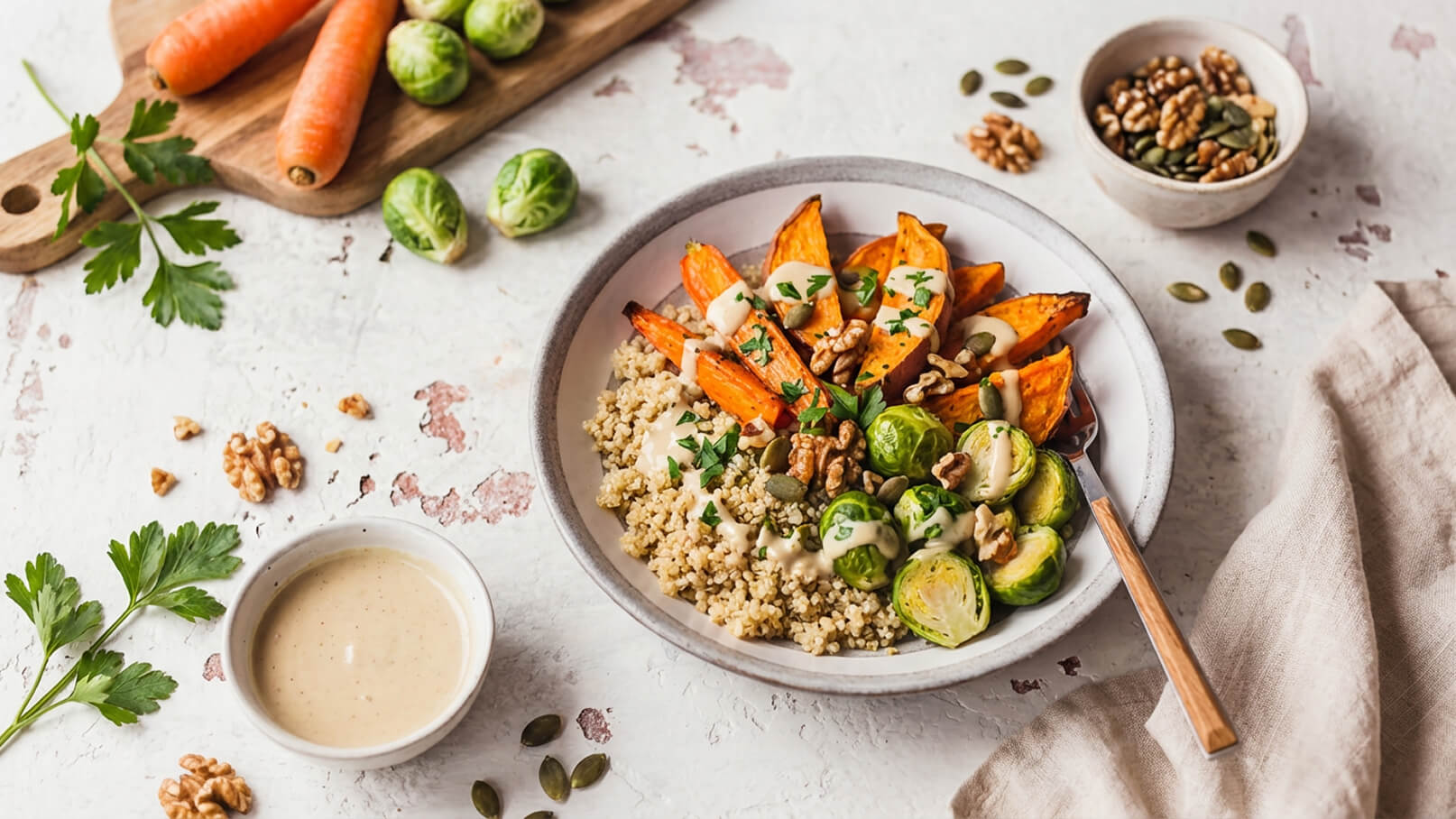 Eine Schüssel mit Quinoa als Basis, garniert mit gerösteten Süßkartoffeln, rohem Rosenkohl und Walnüssen. Darüber befindet sich eine cremige Sauce. Im Hintergrund sind frische Karotten und einige Kürbiskerne zu sehen.
