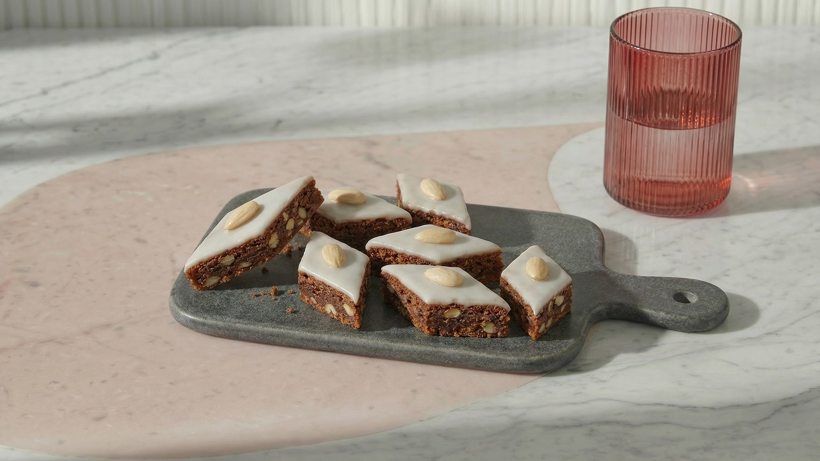 Auf einem grauen Teller liegen mehrere Stücke einer süßen Leckerei mit einer weißen Zuckerglasur und einer ganzen Mandel obendrauf. Der Teller befindet sich auf einer marmorierten Tischfläche. Rechts steht ein pinkfarbener Getränke-Glas.