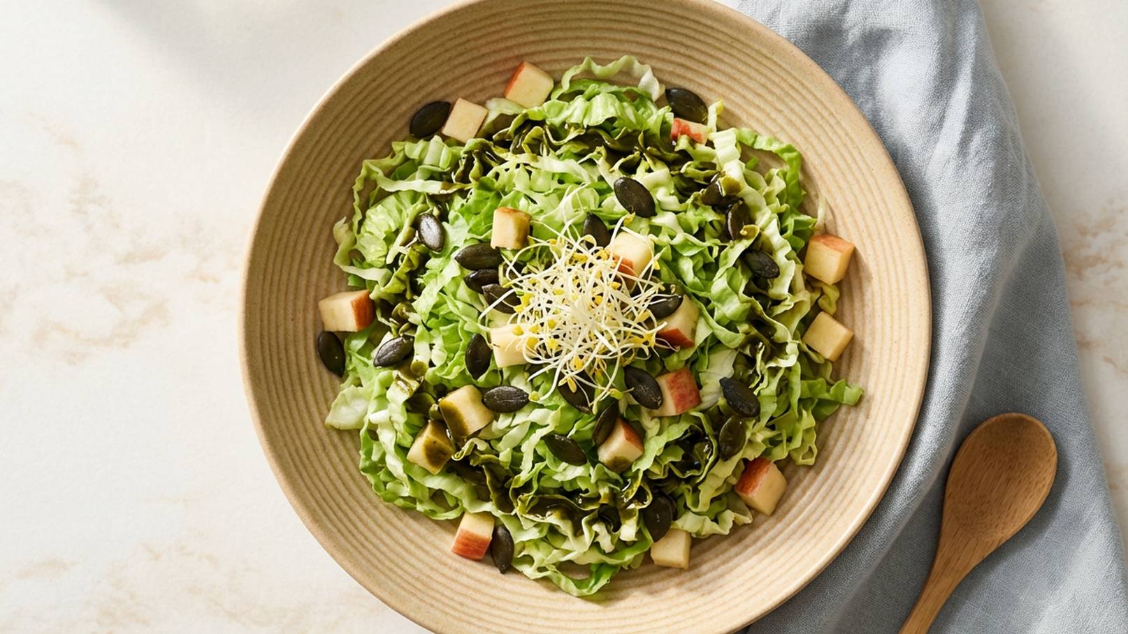 Eine Schüssel gefüllt mit grünem Blattsalat, geschnittenen Äpfeln und Kürbiskernen. Oben auf dem Salat befinden sich Sprossen. Die Schüssel ist hellbraun und steht auf einem hellen Tisch. Eine hölzerne Löffel liegt daneben.